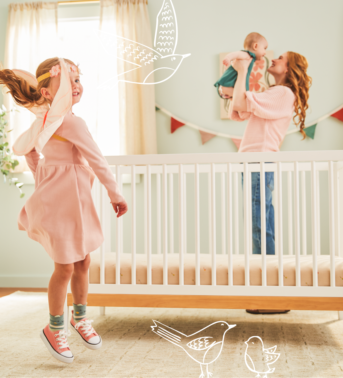Woman holding a baby and standing next to a crib with a young girl in a pink dress in a nursery.